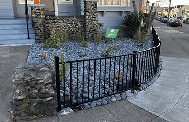 Black metal fence enclosing a rock garden with decorative plants and a green sign.